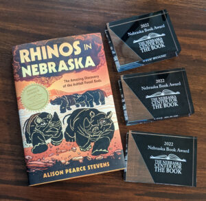 Cover of Rhinos in Nebraska with award winner sticker next to three Nebraska Book Award plaques