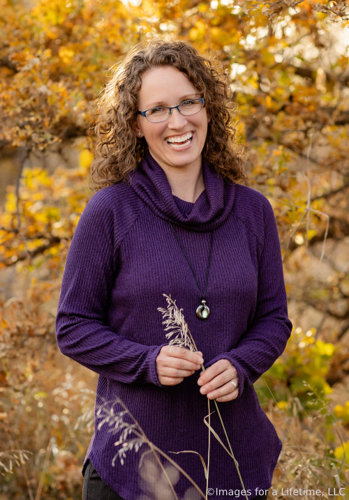 Laughing woman with brown, curly hair wearing glasses and a purple cowl-neck sweater.